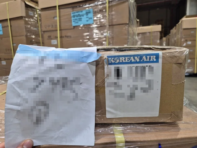 손으로 종이를 들고 있고 옆에 테이프로 포장된 상자가 놓여 있으며 뒤로 상자들이 쌓인 창고가 보이는 모습